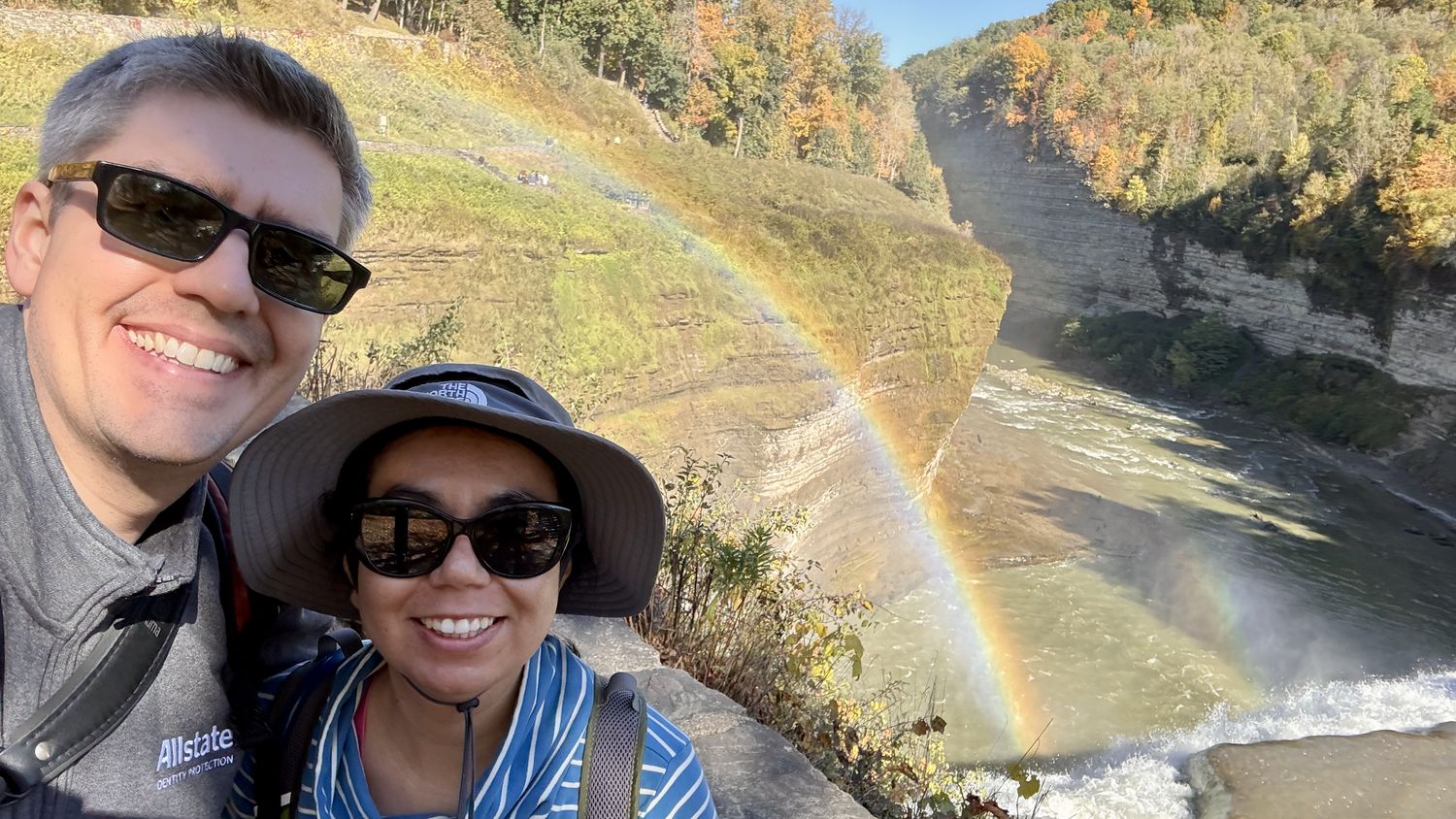 A selfie of Eric and Rebecca, smiling and wearing sunglasses. They are near the top of a gorge and a waterfall, framed by a rainbow.